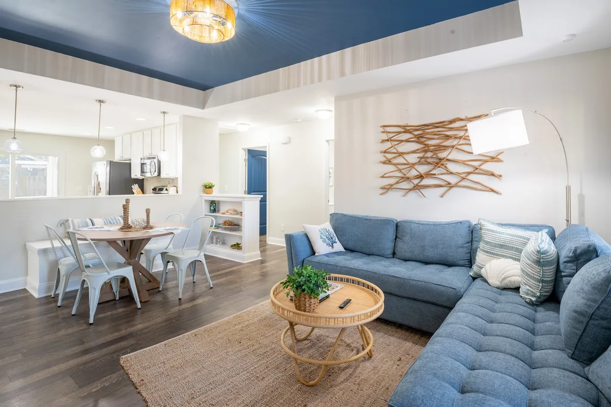 An open, inviting living and dining area with a blue sectional sofa, wooden dining table, woven wall decor, and a round coffee table. The layout is designed for group gatherings and relaxed socializing, with a cozy and welcoming ambiance.