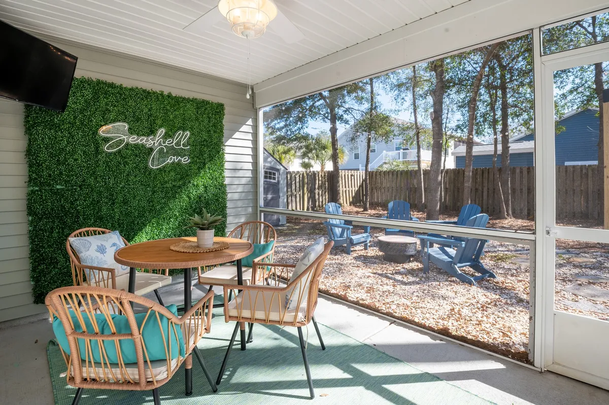 The hero image showcases a stylish interior with a vibrant green accent wall adorned with a 'Seashell Cove' sign, a welcoming dining table surrounded by chairs, and a view of the inviting outdoor seating area, all bathed in natural light. The setup hints at cheerful gatherings, cozy meals, and seamless indoor-outdoor living, perfect for making vacation memories with loved ones and pets.