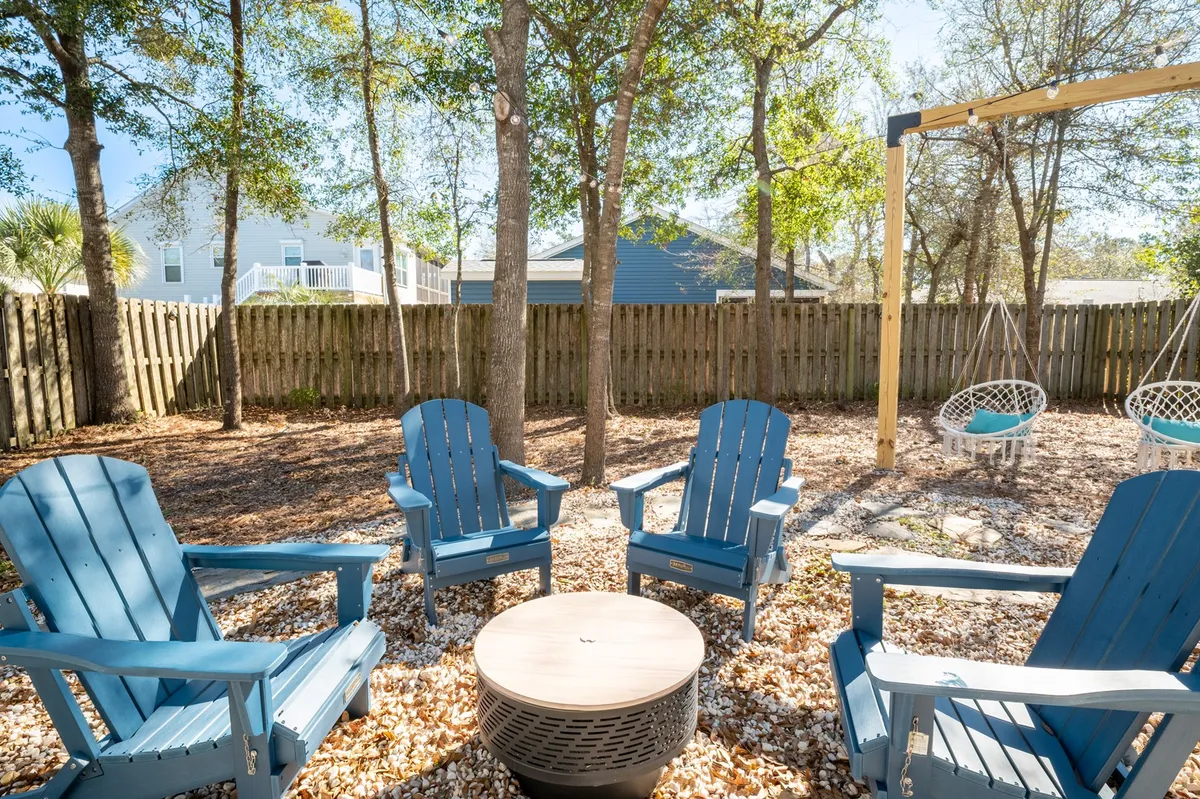 The image showcases a vibrant backyard scene featuring blue Adirondack chairs arranged around a charcoal fire pit with hanging swings nearby. The setting is casual and inviting, ideal for both daytime relaxation and evening gatherings under the stars. The lush, fenced backdrop provides a sense of privacy and tranquility.