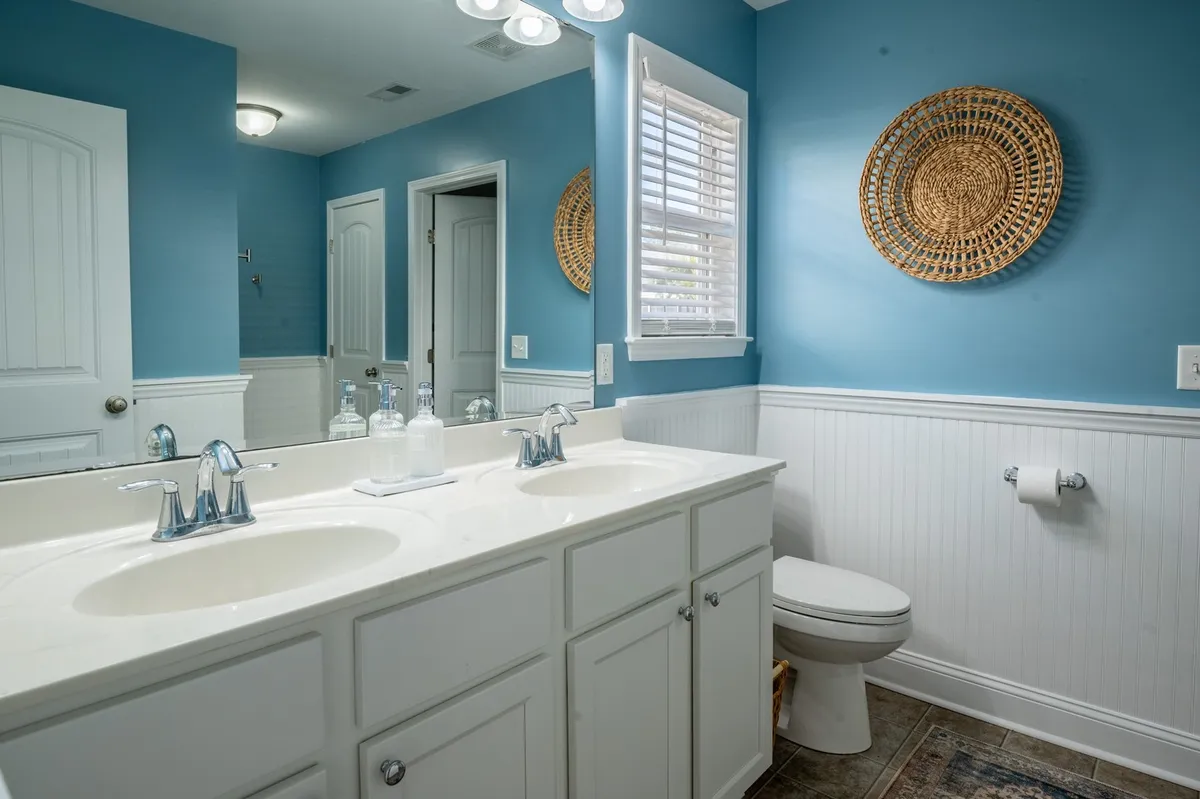 An inviting bathroom with a double vanity featuring two sinks, blue walls, and woven wall decor. The space feels spacious and well-suited for shared use, reflecting comfort and modern style.