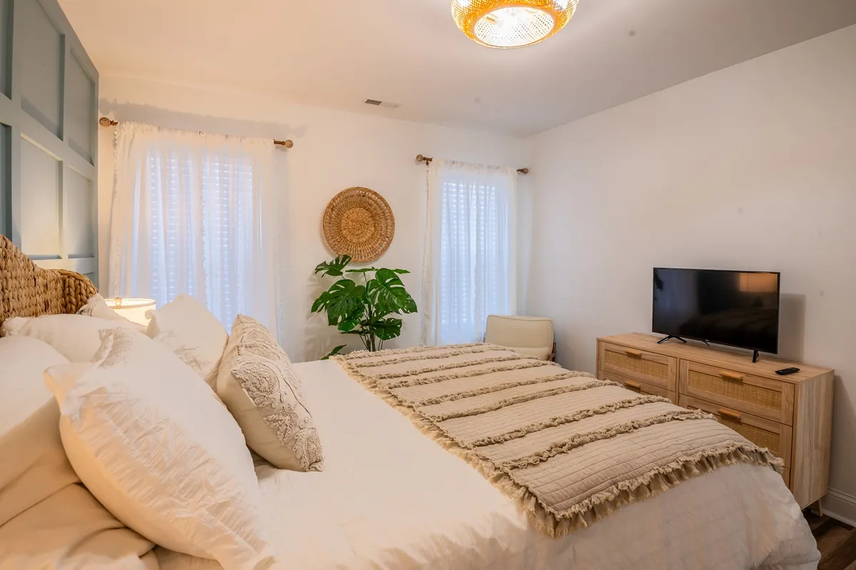 A cozy bedroom showcases a bed with decorative pillows, woven wall decor, an indoor plant, and a television. The overall mood is calm and inviting, perfect for unwinding after a day of Oak Island adventures.