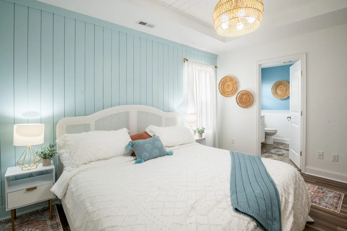 A queen bedroom showcasing a bed with decorative pillows, stylish bedside lamps, woven wall decor, and a blue accent wall. The space is cozy, well-lit, and exudes a sense of calm and sophistication.