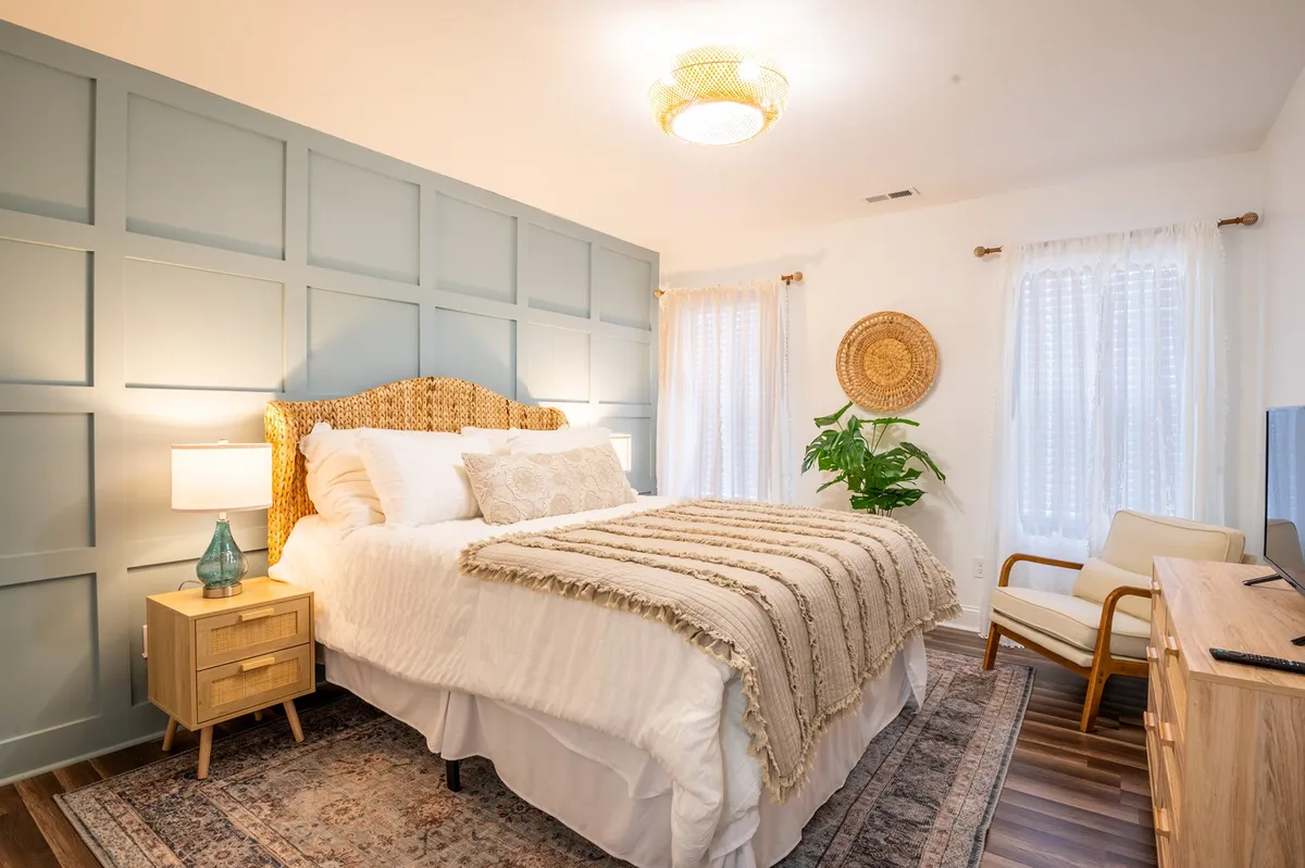 A serene bedroom featuring a decorative headboard, textured throw blanket, decorative wall paneling, large windows with sheer curtains, an indoor plant, and a nightstand with a lamp. Soft natural light fills the space, creating a restful and inviting atmosphere.