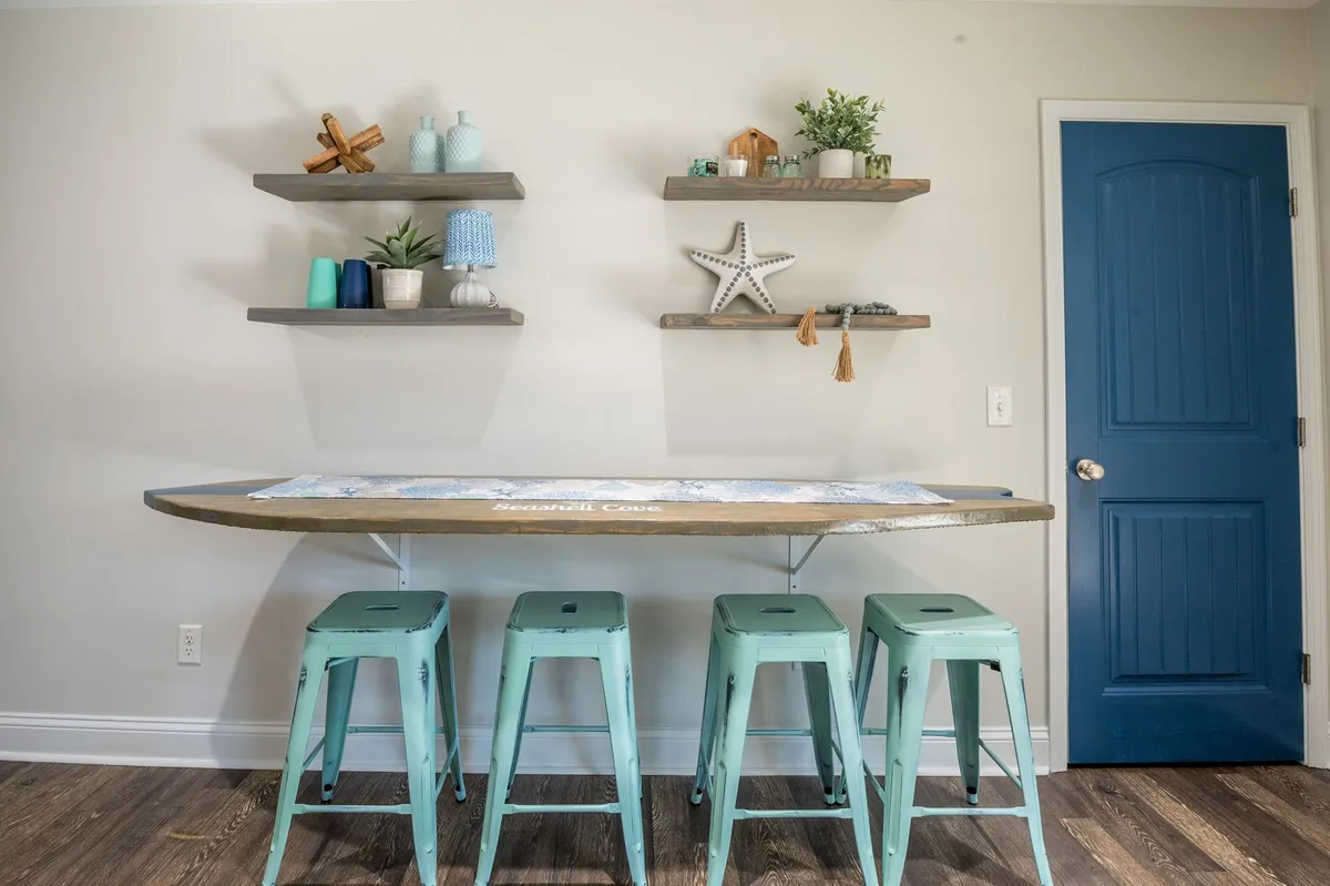 A playful interior shot featuring a surfboard-themed table, blue door, and decorative shelving. The mix of beachy decor and relaxed seating hints at casual meals and lively social gatherings, echoing the property’s coastal personality.