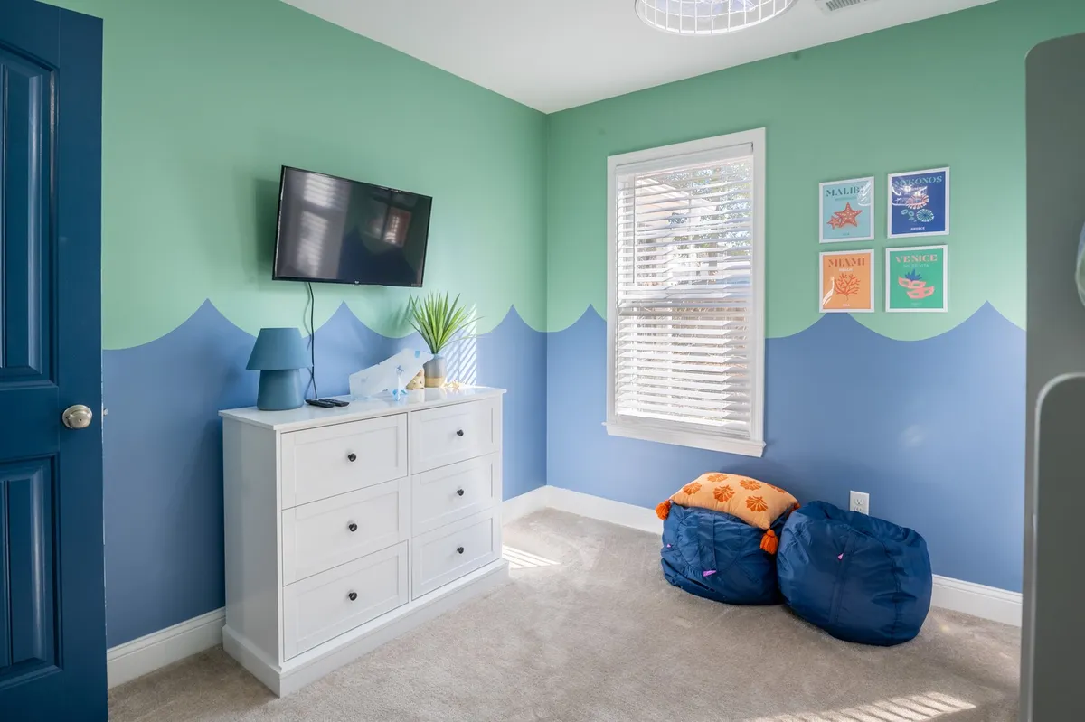 A playful bunkroom featuring bean bag chairs, a colorful wall design, a white dresser, television, and decorative prints. The space is fun, inviting, and perfect for children’s activities, reading, or movie nights.