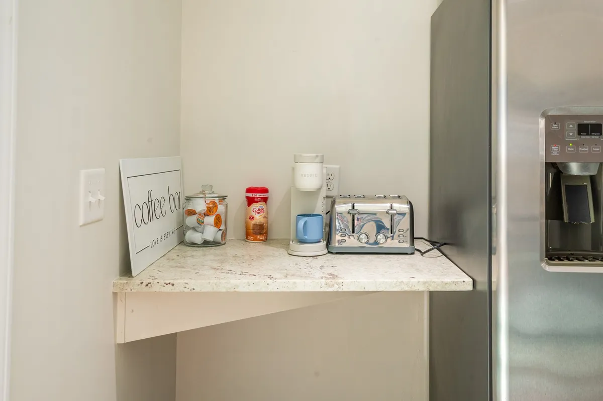 A cozy coffee station featuring a coffee bar sign, coffee maker, toaster, and neatly arranged sugar and cream containers. The setup exudes warmth and hospitality, inviting guests to enjoy a slow morning or chat over coffee.